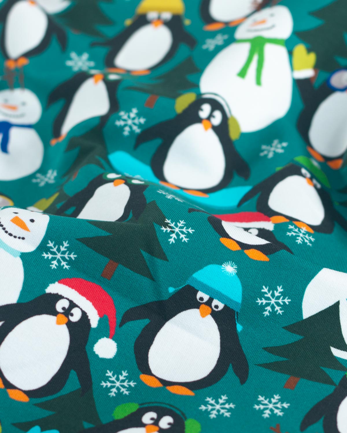 A close-up of a festive cotton jersey fabric featuring a repeating pattern of cartoon penguins, snowmen, snowflakes, and Christmas trees on a teal background.