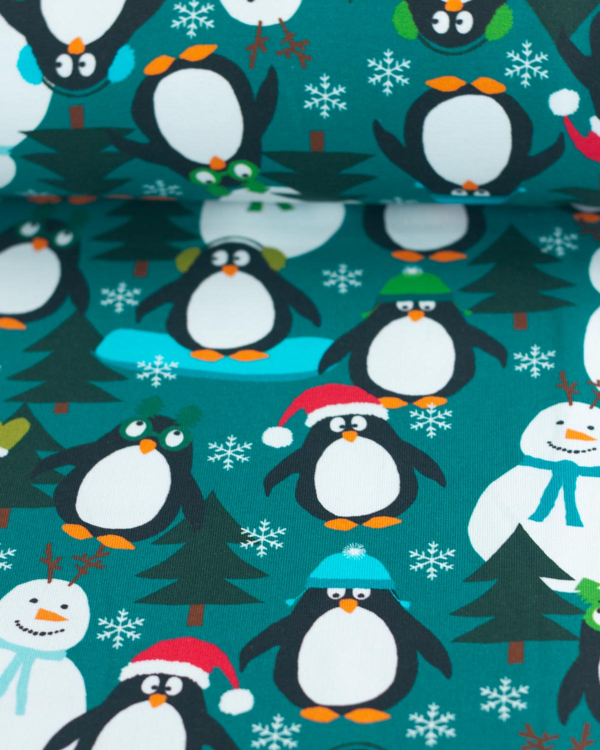 A close-up of a festive jersey fabric featuring a repeating pattern of cartoon penguins, snowmen, Christmas trees, and snowflakes on a teal background.