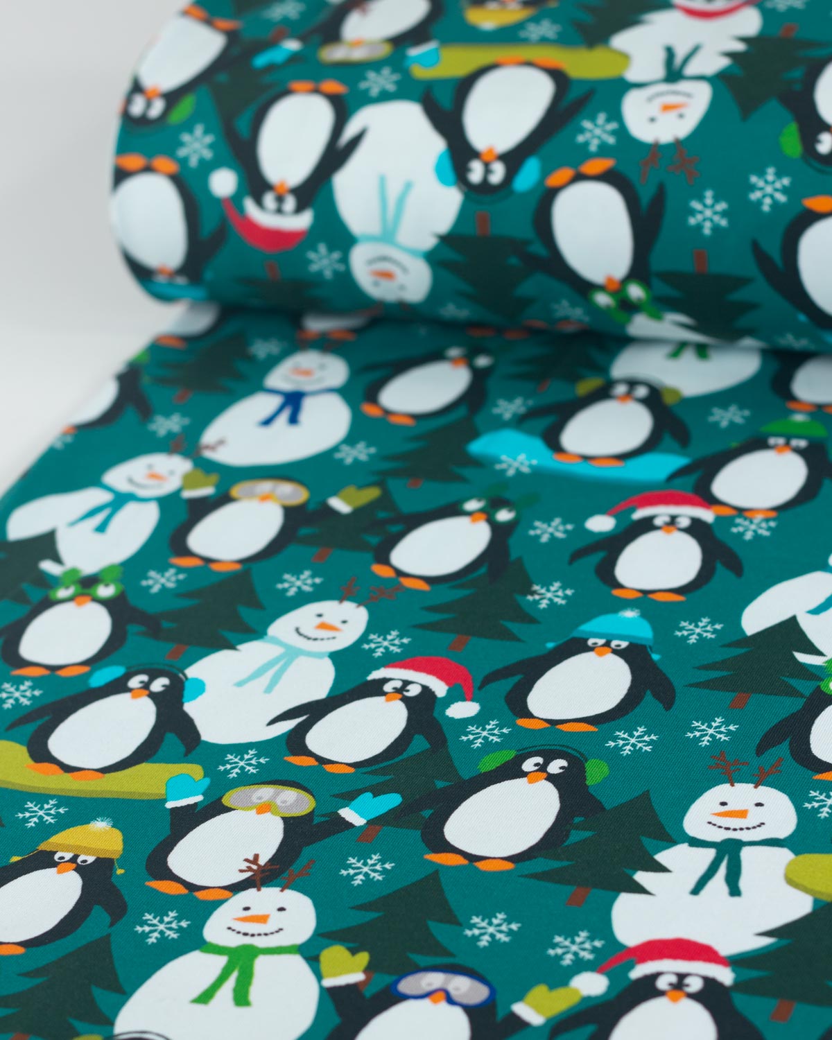 A close-up of a festive cotton jersey fabric featuring a repeating pattern of cartoon penguins, snowmen, snow trees, and snowflakes on a teal background.