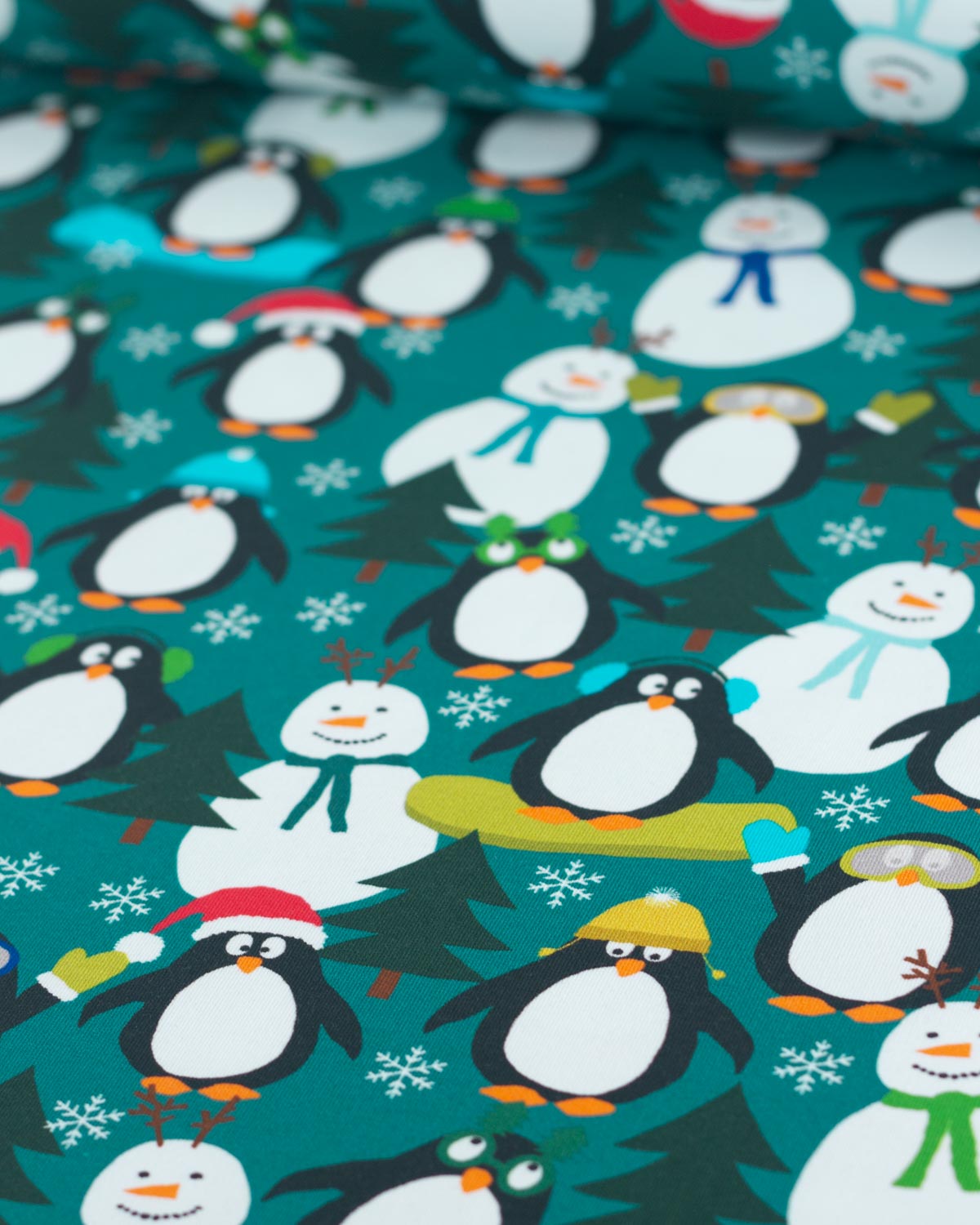 A close-up of  Christmas cotton jersey fabric featuring cartoon penguins, snowmen, snow trees, and snowflakes on a teal background.