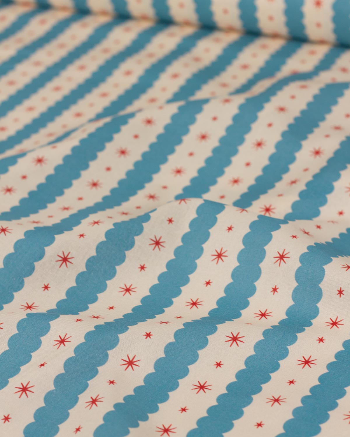 A close-up of patterned cotton fabric with diagonal blue scalloped stripes on an off-white background, accented with small red starburst motifs.