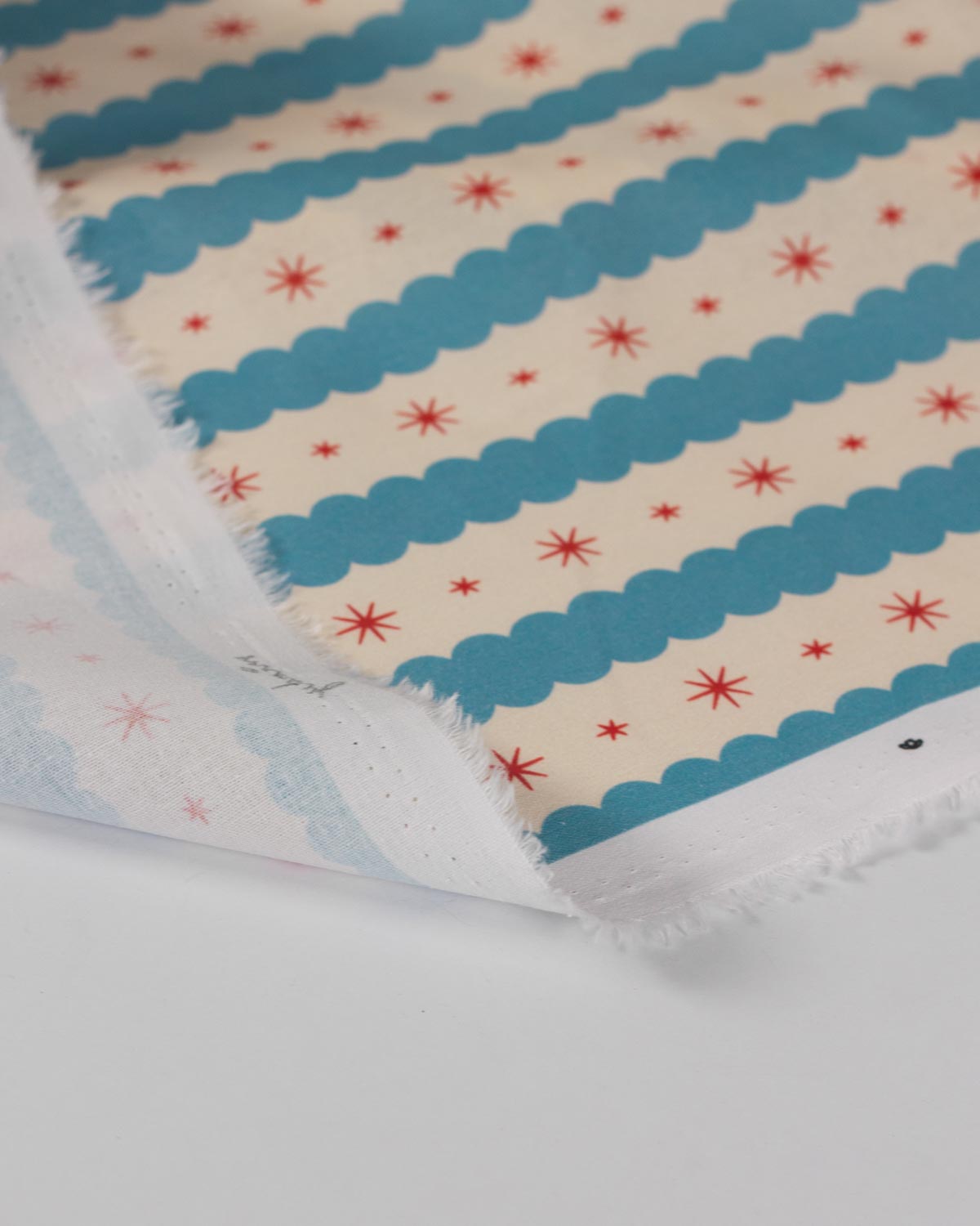 A close-up of patterned fabric with alternating blue scalloped stripes and red starburst motifs on a cream background, folded over to show the selvedge.