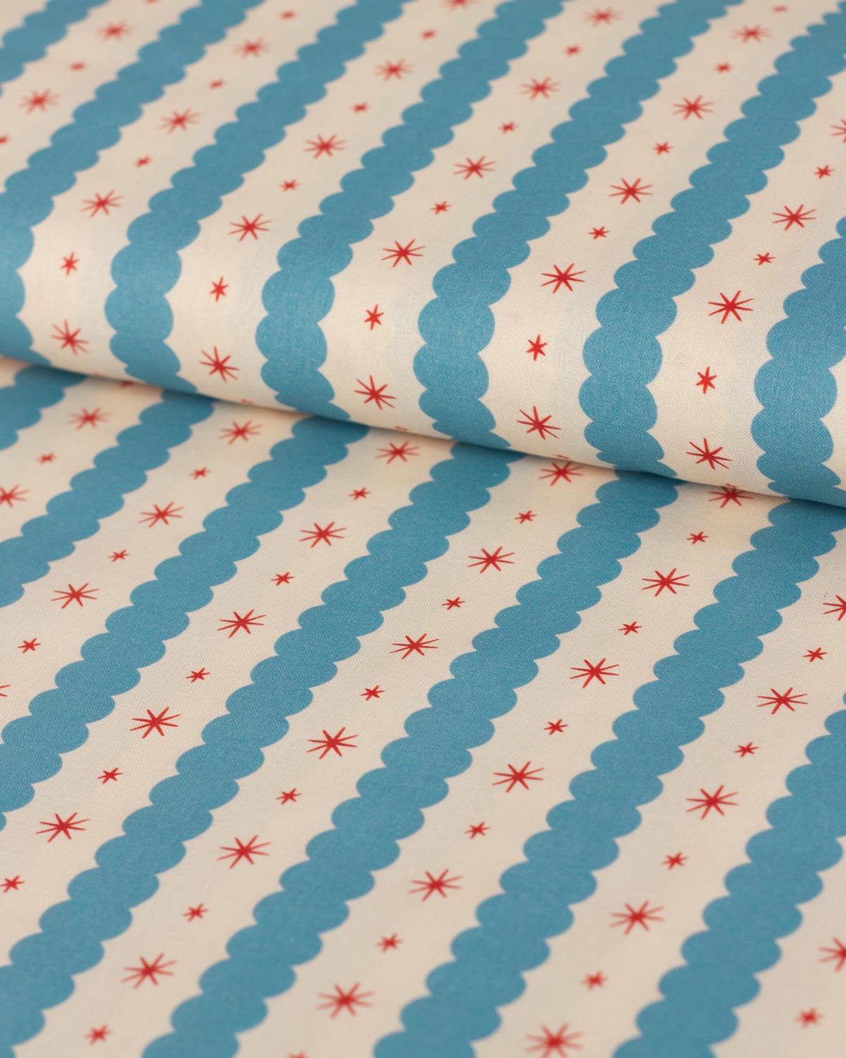 A close-up of patterned fabric with diagonal blue scalloped stripes on an off-white background, decorated with small red starburst motifs.