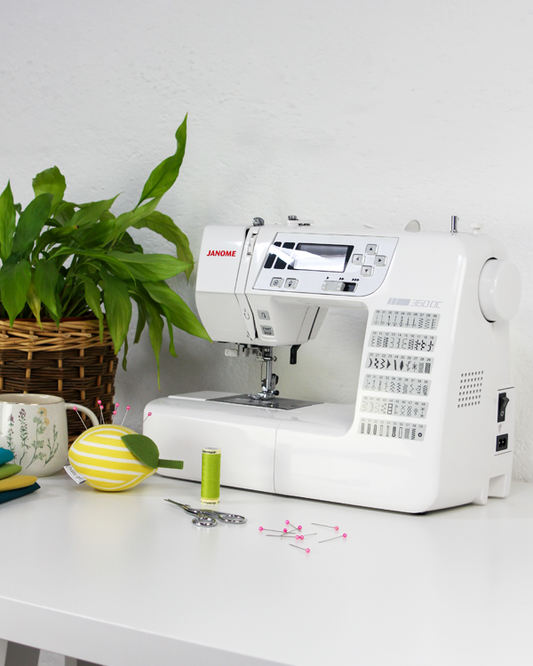 White Janome sewing machine on a white table with a plant and sewing accessories.
