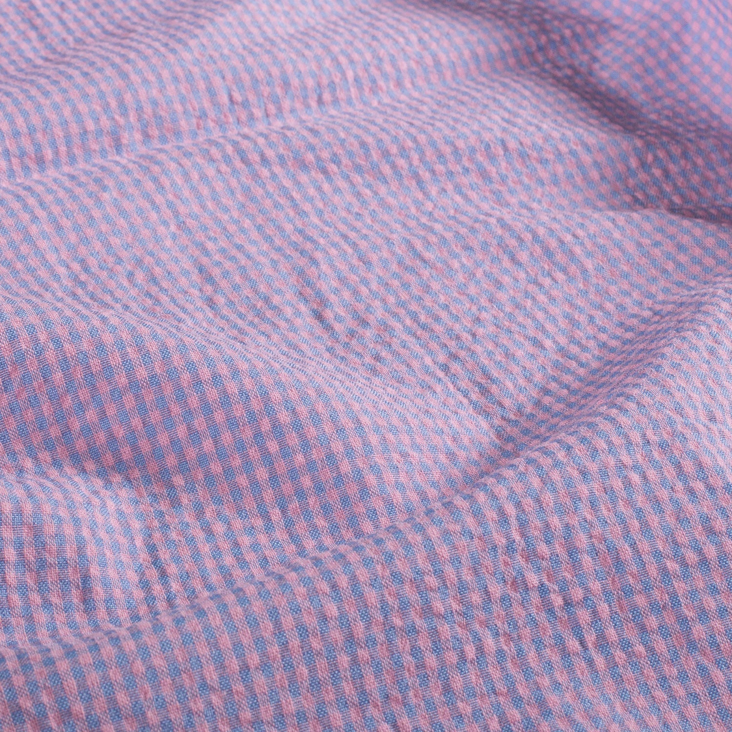 A close-up of a textured fabric with a fine pink and blue gingham check pattern, showing soft folds and shadows.