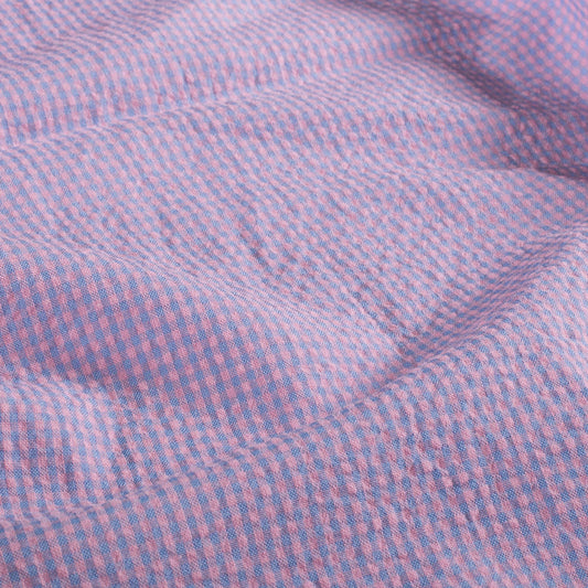 A close-up of a textured fabric with a fine pink and blue gingham check pattern, showing soft folds and shadows.