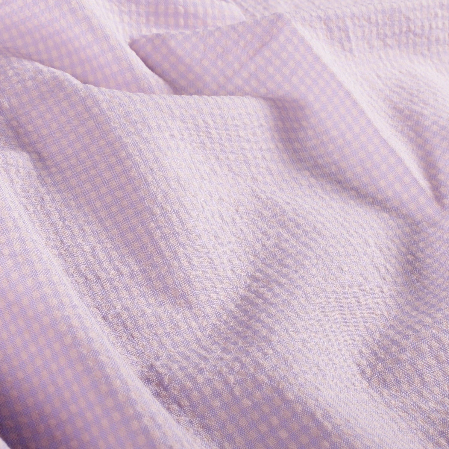 A close-up of a light lavender and white gingham patterned fabric with a textured, waffle-like weave, shown in soft folds.