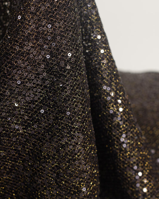 Close-up of a black fishnet tulle fabric with shimmering gold lurex and black reflective sequins, draped in soft folds against a light neutral background.