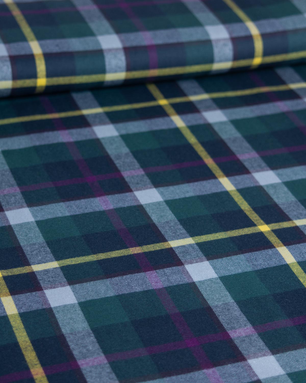 A close-up of plaid cotton fabric with a tartan pattern in dark green, navy, light gray, and thin yellow and purple stripes.