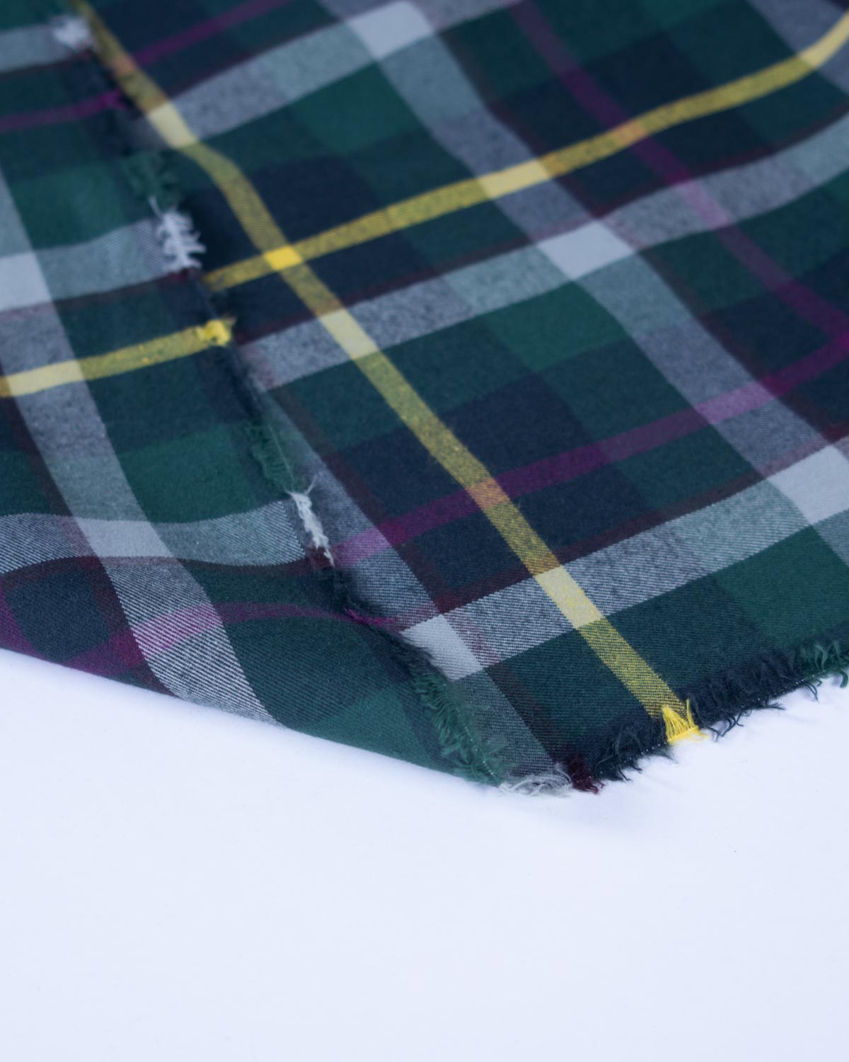 A close-up of a folded plaid fabric with a tartan pattern in dark green, navy, purple, gray, and yellow, showing a frayed edge against a plain white background.