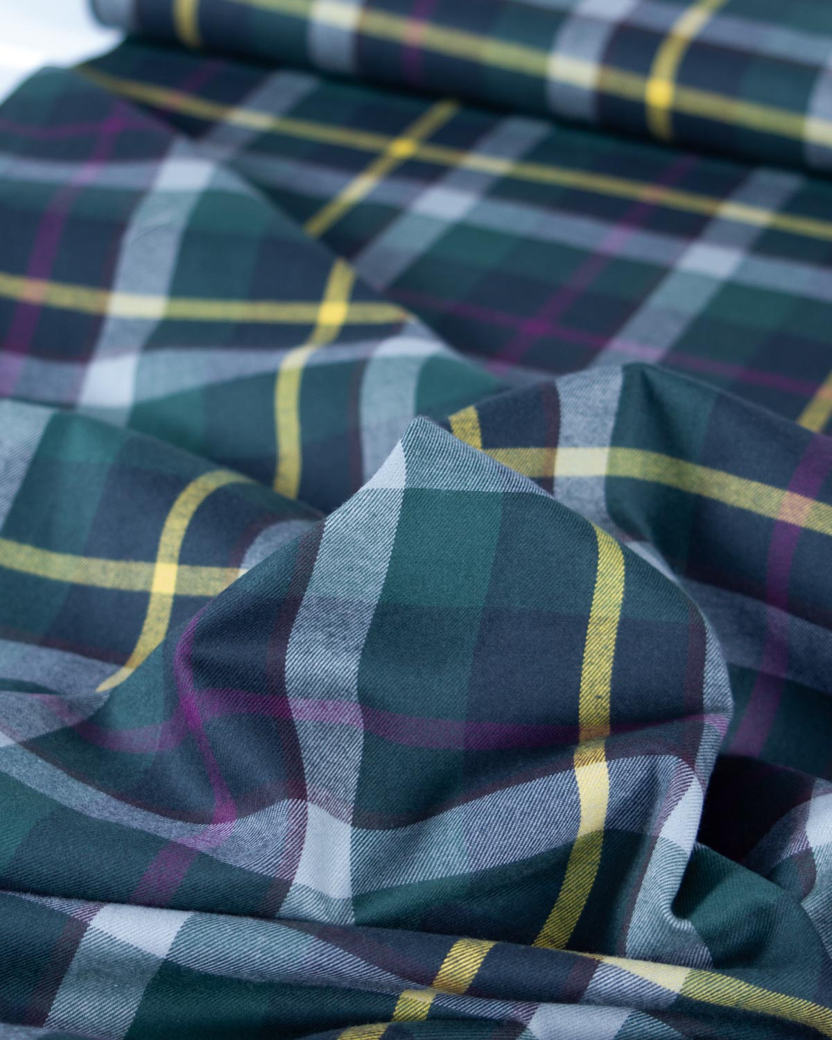 A close-up of draped tartan cotton fabric with a plaid pattern in dark green, slate gray, purple, and yellow.