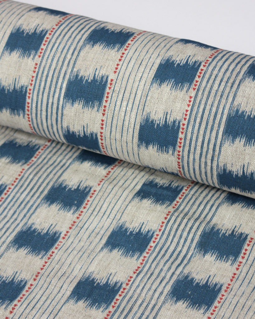 A close-up of a rolled piece of upholstery fabric with a blue and cream ikat pattern, accented by thin red dotted vertical lines.
