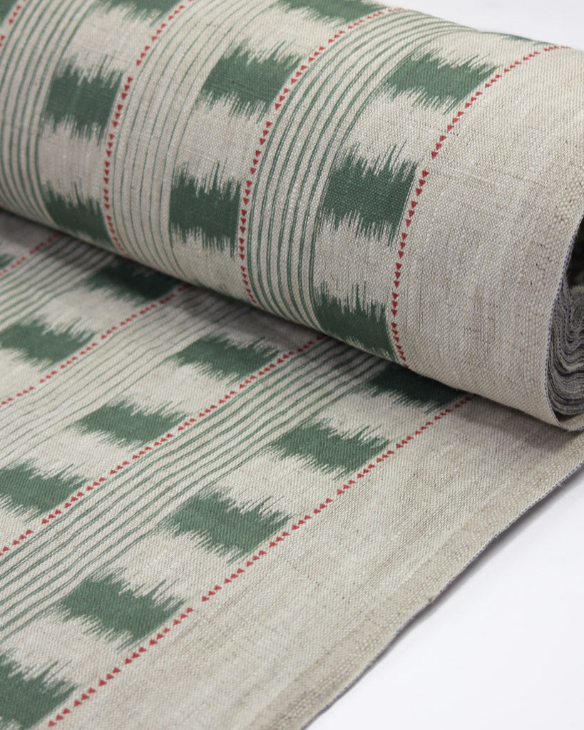 A close-up of a rolled woven textile, likely a rug or fabric panel, featuring a green and beige ikat-style pattern with thin red dotted accent lines 