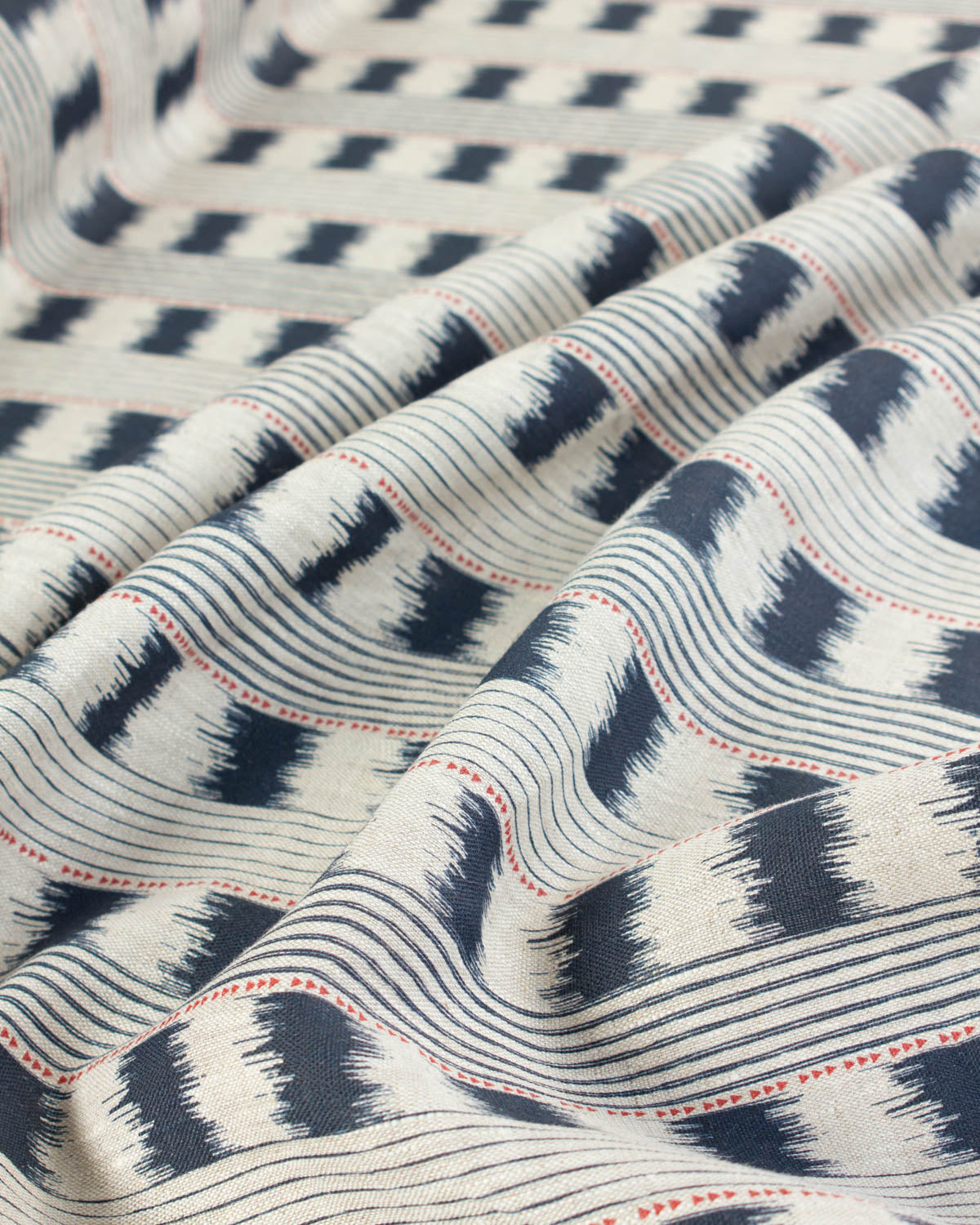 A close-up of draped upholstery fabric with a cream base and navy geometric ikat-style patterns, accented by thin red dotted lines.