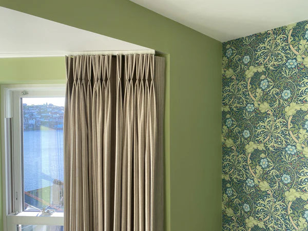Beige pinch pleat made to measure curtains in front of a window with green walls and floral William morris wallpaper.