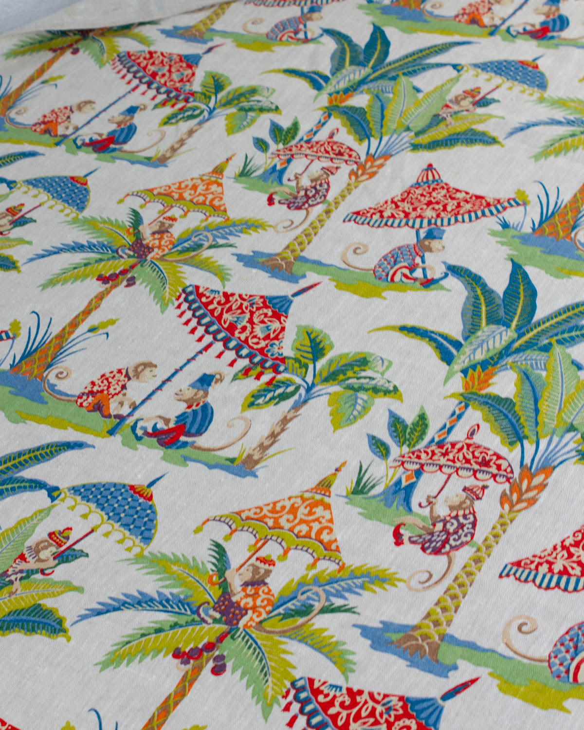 A close-up of a printed fabric featuring a tropical beach scene with palm trees, beach umbrellas, and monkey figures in bright colours on a linen upholstery fabric