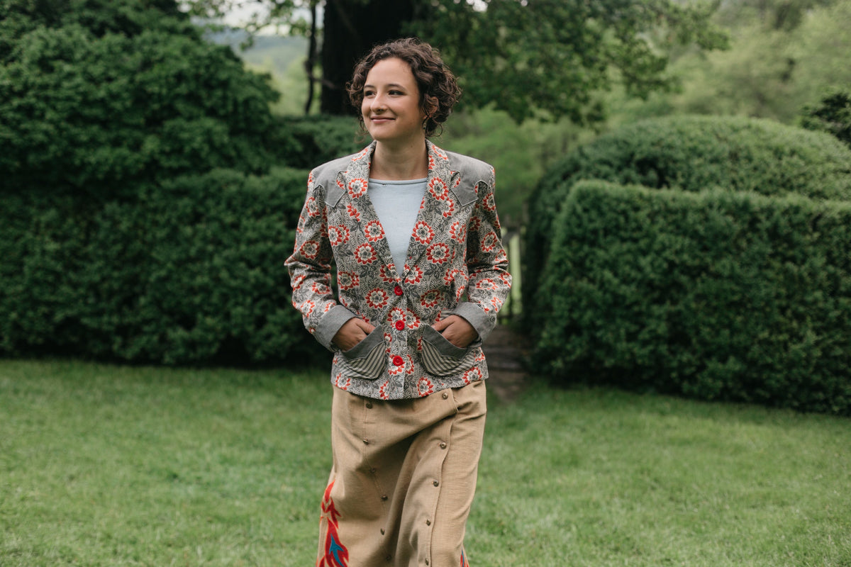 Young brunette woman smiling walking surrounded by greenery wearing the 242 Rodeo Cowgirl Jacket with hands in the pockets.