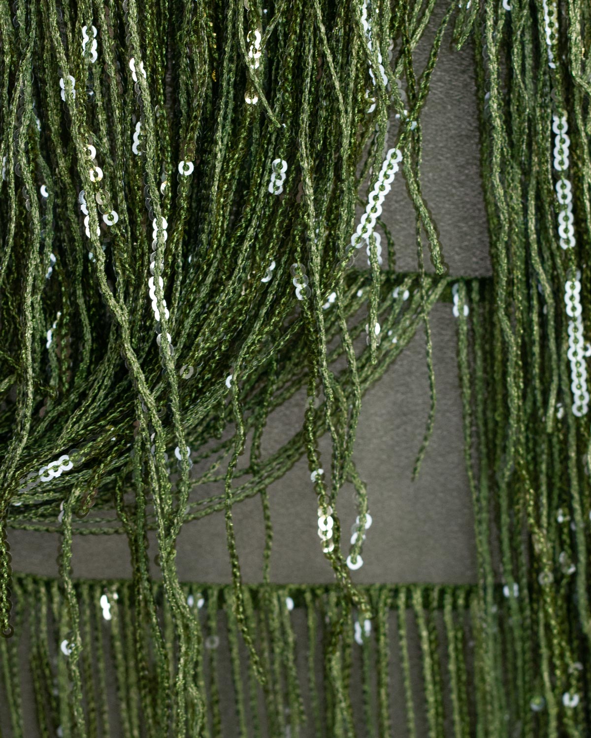 Close-up of a dark olive green fringe polyester fabric with shimmering silver sequined tassels hanging against a olive green fabric backing.
