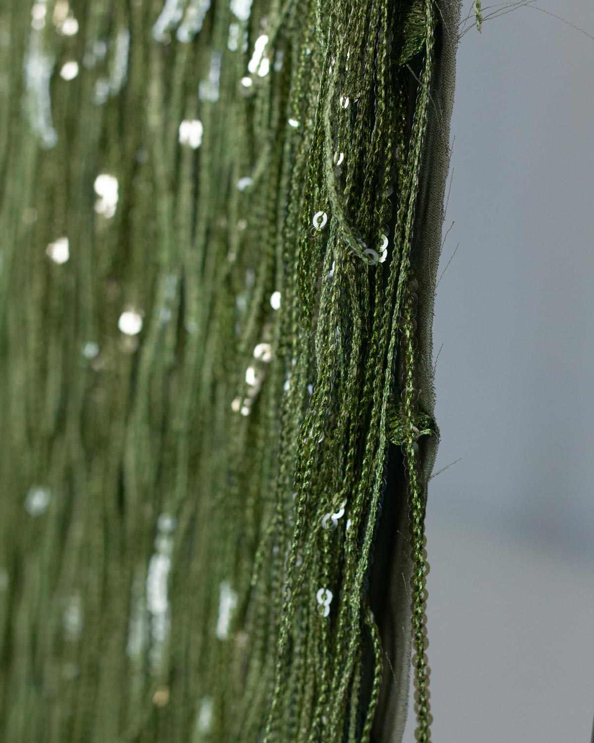 Close-up of olive green sequined fringe tassel polyester fabric hanging vertically, with small reflective sequins catching the light against a soft white background.