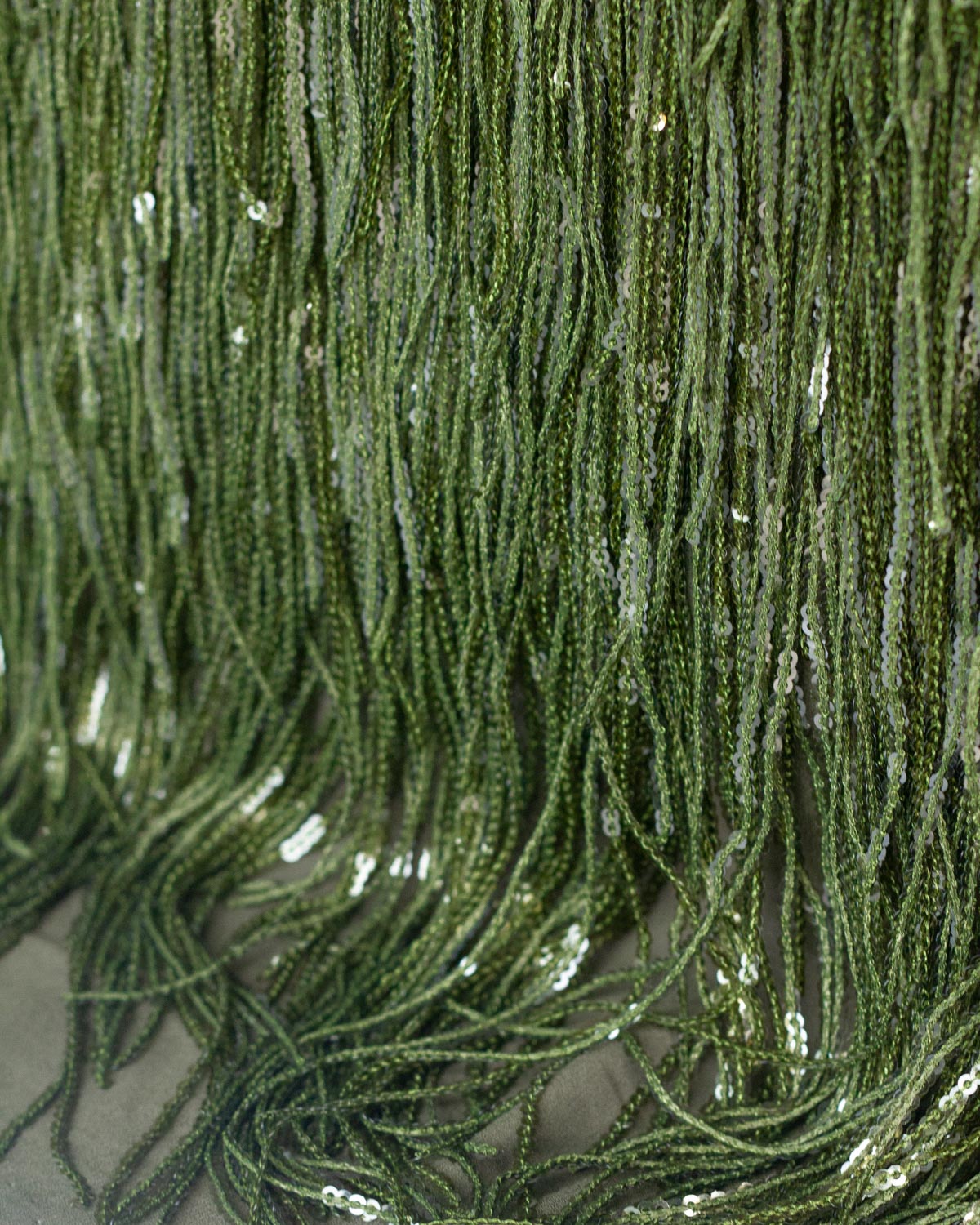 Close-up of dark green sequined fringe polyester fabric with shimmering reflective sequins hanging vertically.