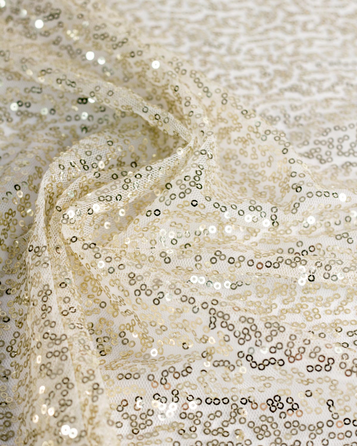 Close-up of a sheer champagne-colored sequin tulle fabric with shimmering gold metallic sequins and a delicate mesh backing, draped in soft folds.