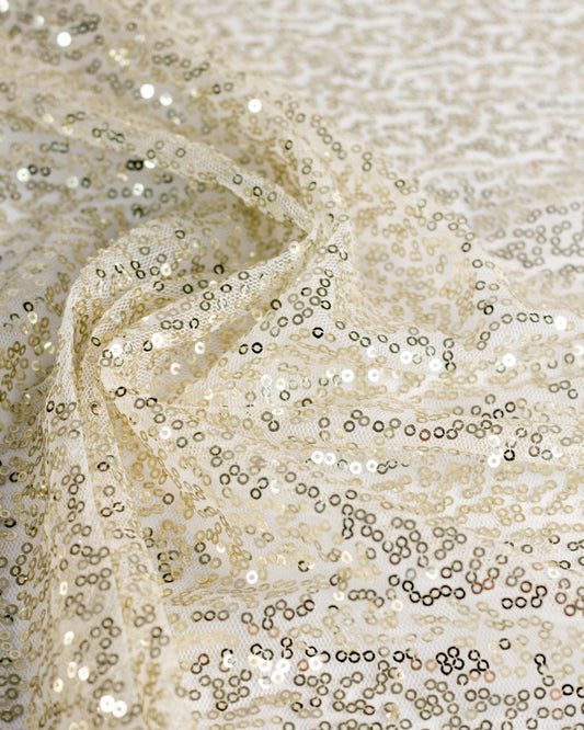 Close-up of a sheer champagne-colored sequin tulle fabric with shimmering gold metallic sequins and a delicate mesh backing, draped in soft folds.