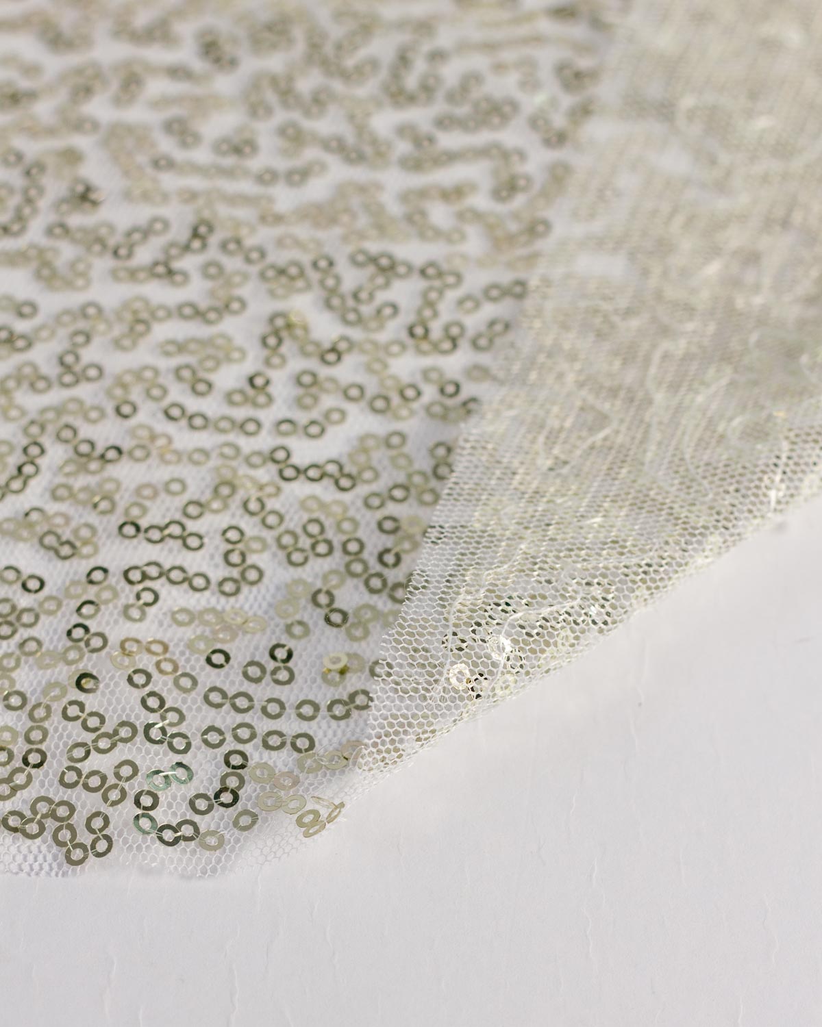 A close-up of a sheer white tulle fabric decorated with dense, small metallic gold champagne sequins, folded diagonally over a plain white surface to show the textured reversed side. 