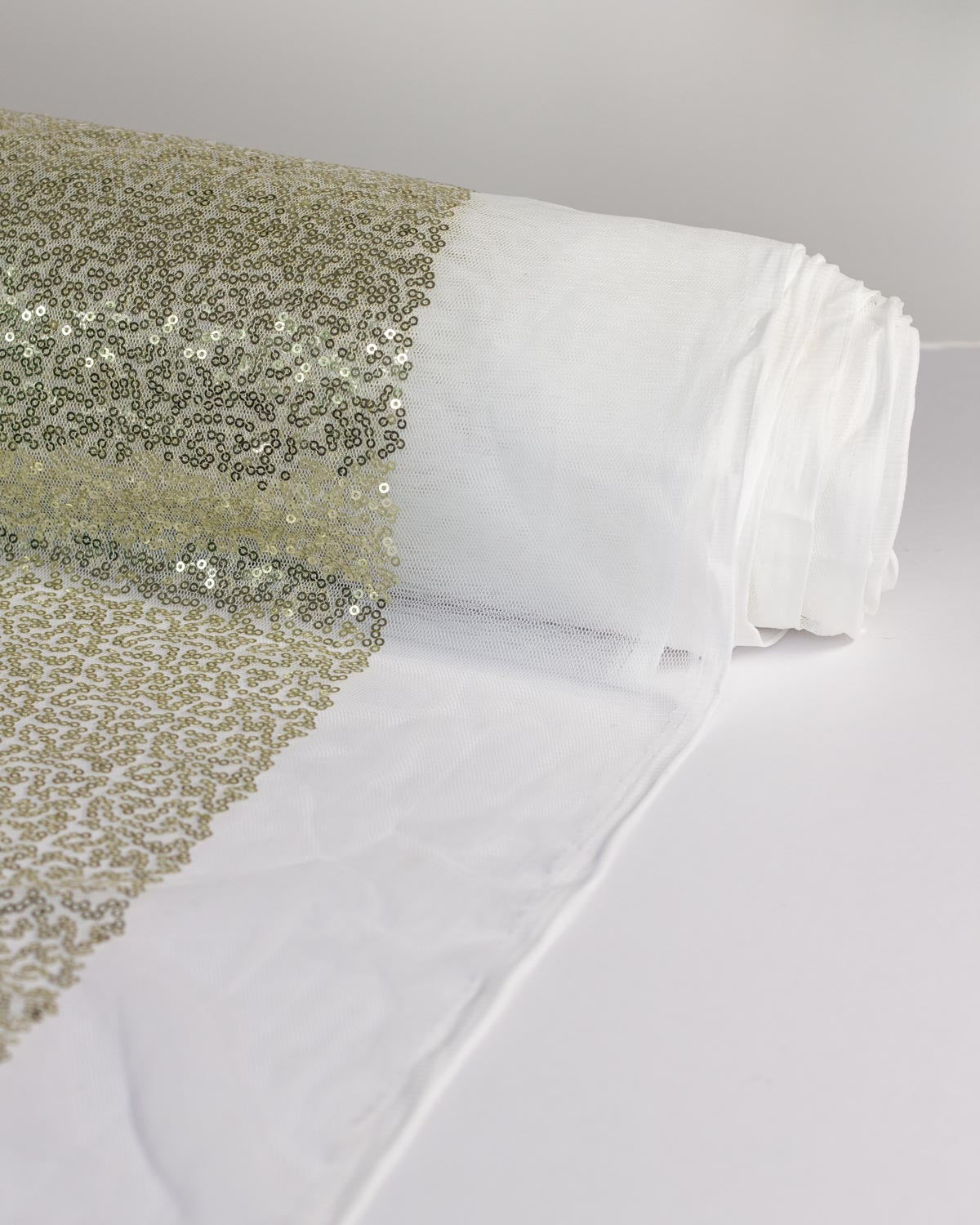 A roll of white tulle fabric with densely packed champagne gold sequins, unrolled on a clean white background.