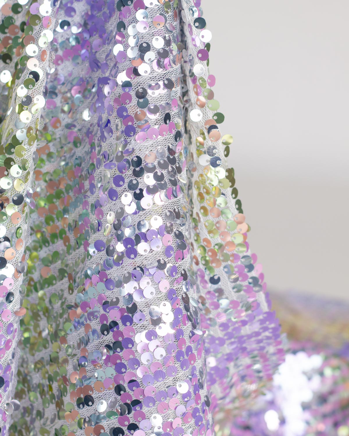 Close-up of a sheer tulle fabric covered in iridescent silver, lavender, pink, gold, yellow and green sequins, draped against a light neutral background.
