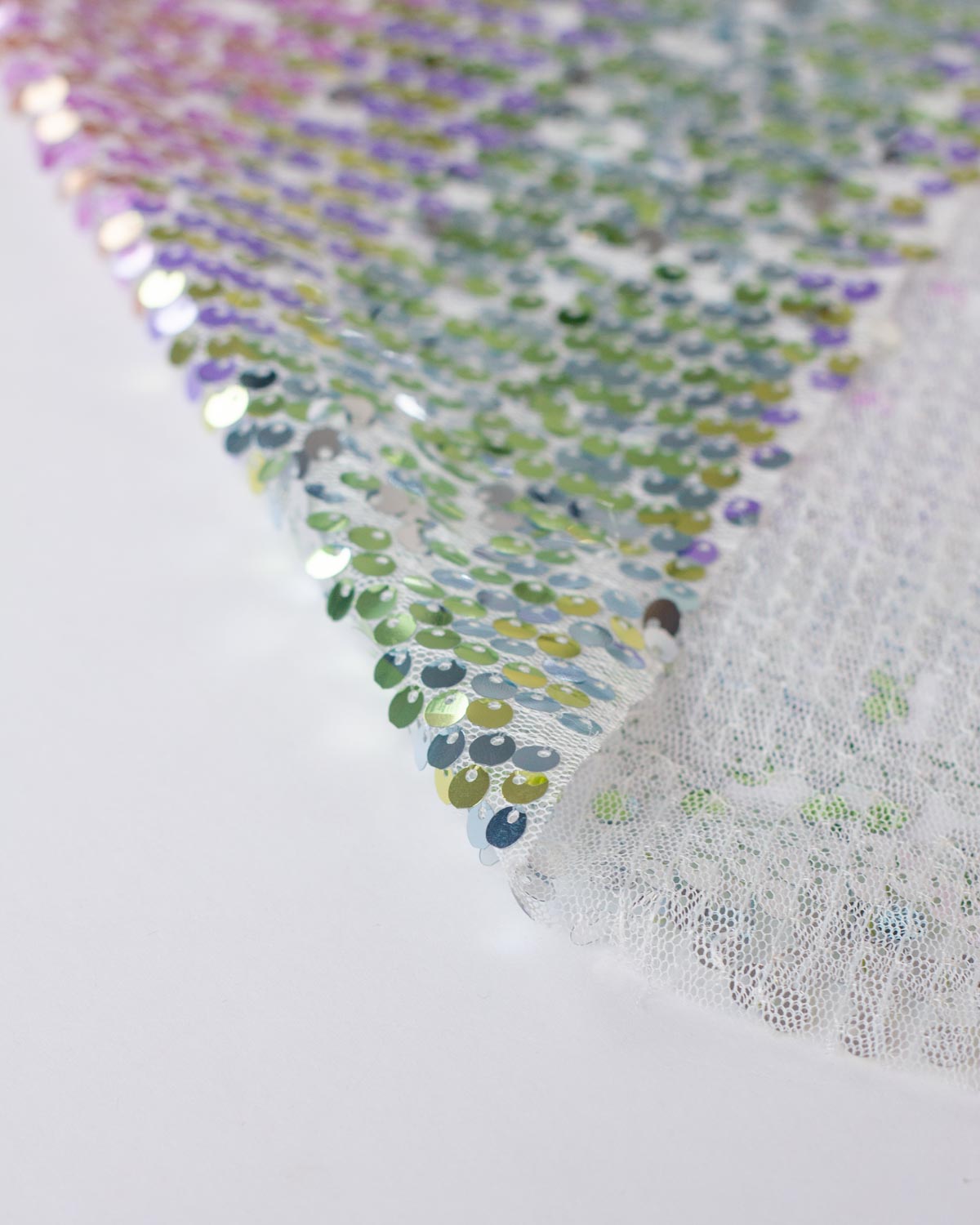 Close-up of a white tulle fabric with iridescent sequins that shift between green, blue, purple, and pink, laid diagonally on a white background showing its reverse side. 