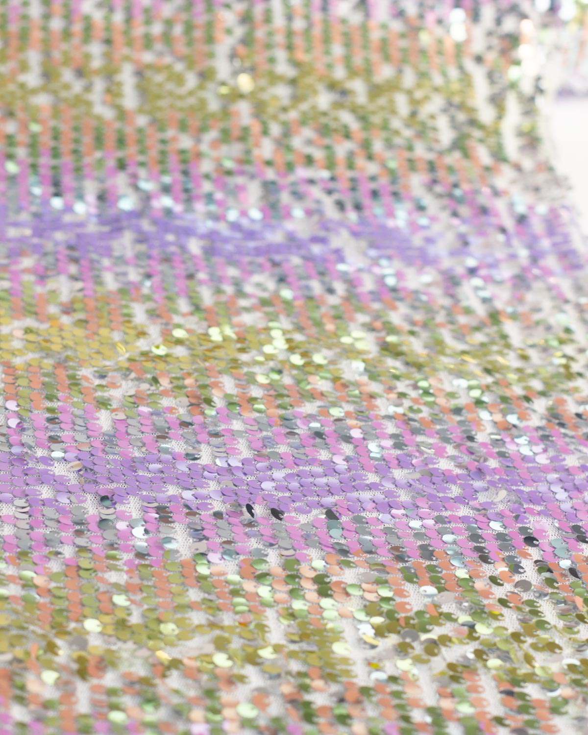 A close-up of a sequined tulle fabric featuring horizontal stripes of lavender purple, soft peach pink, and metallic gold, with shimmering iridescent silver sequins.