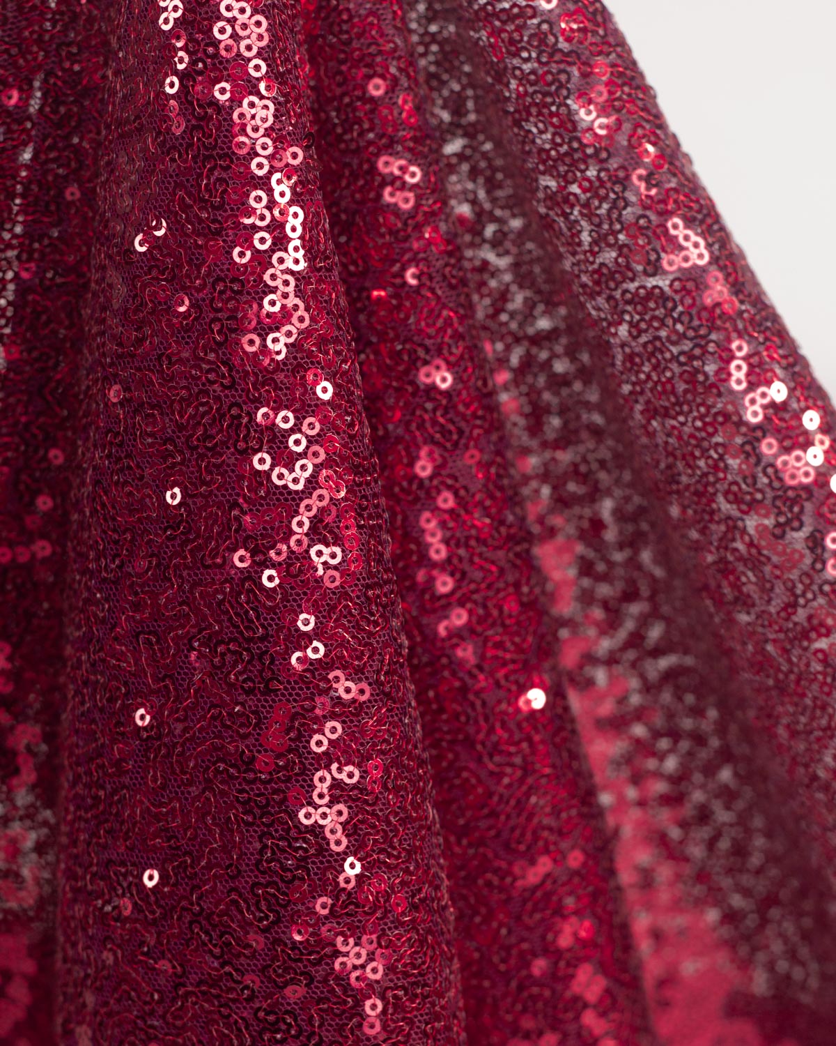 A close-up of draped deep ruby red sequined tulle fabric with dense circular sequins, shown in soft folds against a light gray background.