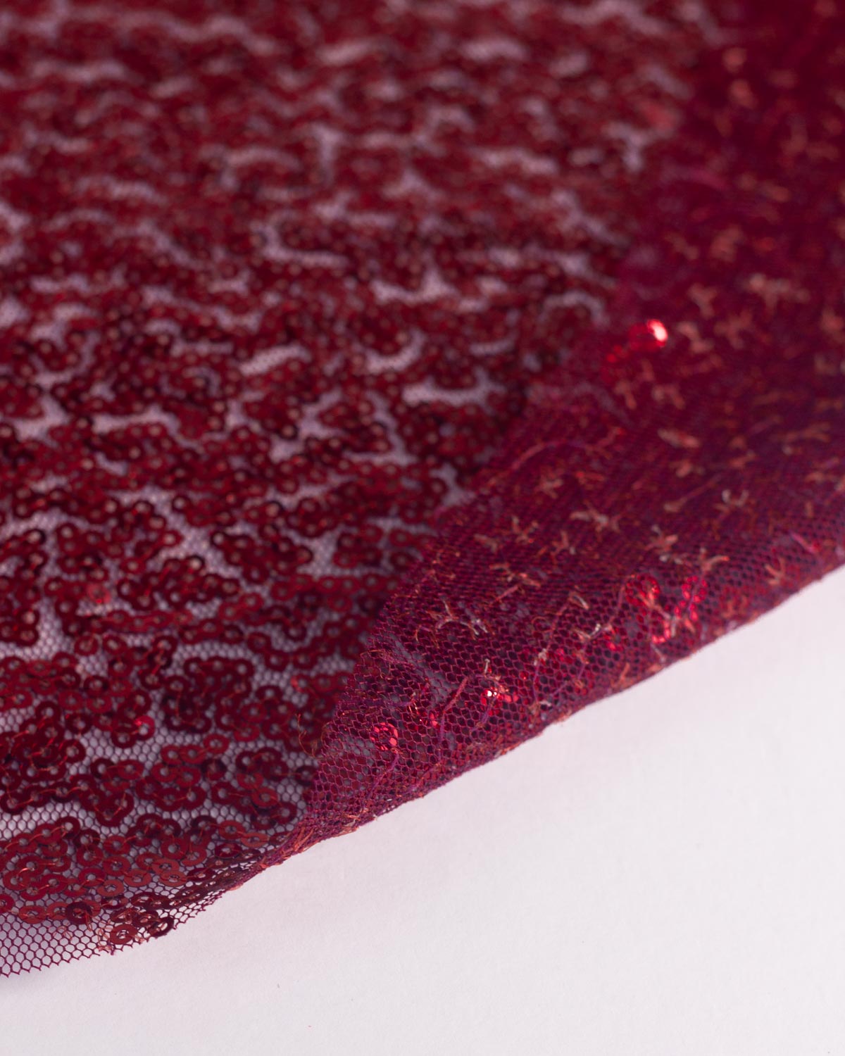 A close-up of deep ruby red sequined tulle fabric draped over a plain white surface, showing the sheer mesh underside and dense sequin pattern.