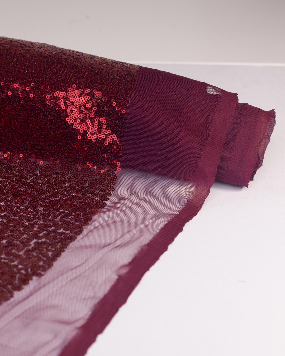 A roll of ruby red sequin polyester tulle fabric, shown partially unrolled on a white surface.