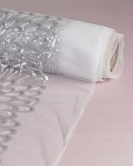 A roll of white polyester tulle fabric featuring silver sequins and white embroidery in feathers or leaf pattern partially unrolled onto a light pink background.