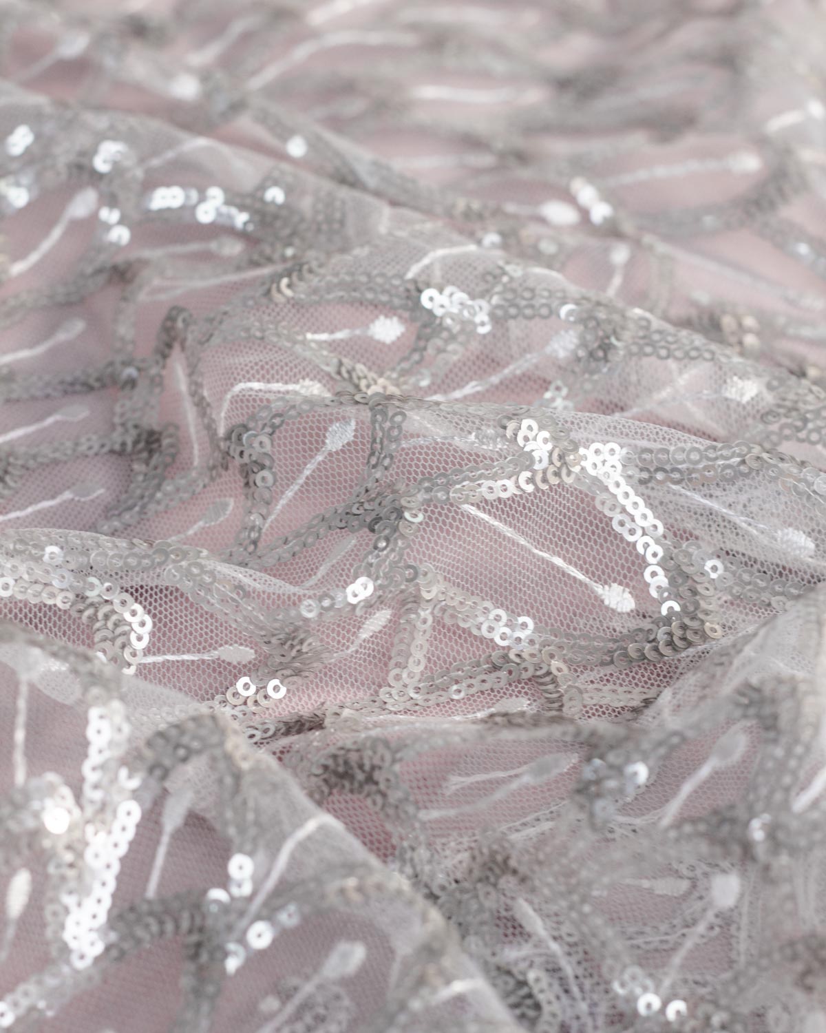 Close-up of sheer white tulle mesh fabric with silver sequins and white embroidery arranged in a feather or leaf pattern over a dusty pink background.