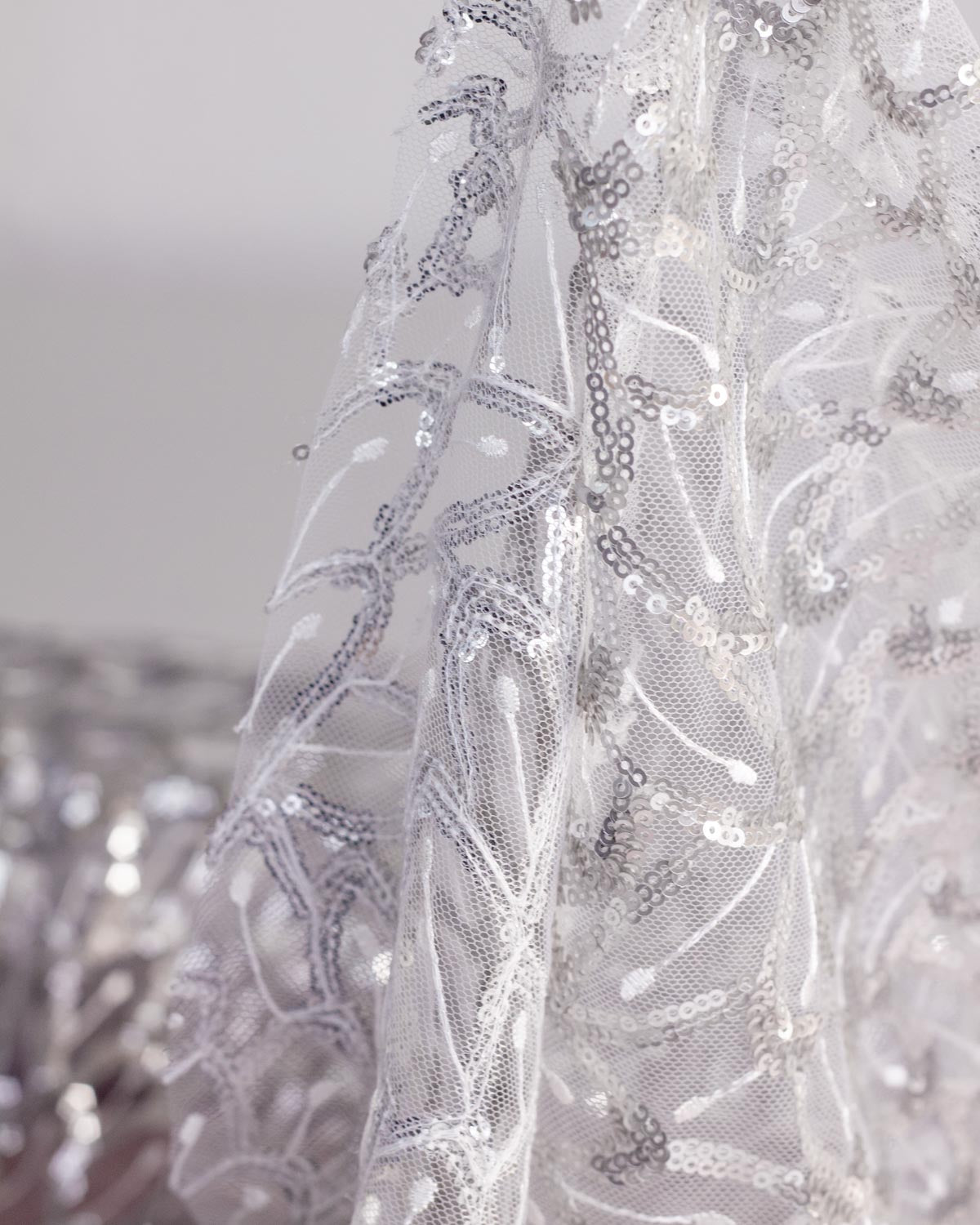 Close-up of draped white sheer polyester tulle mesh fabric with intricate silver sequins and white embroidery in a feather and leaf pattern. 