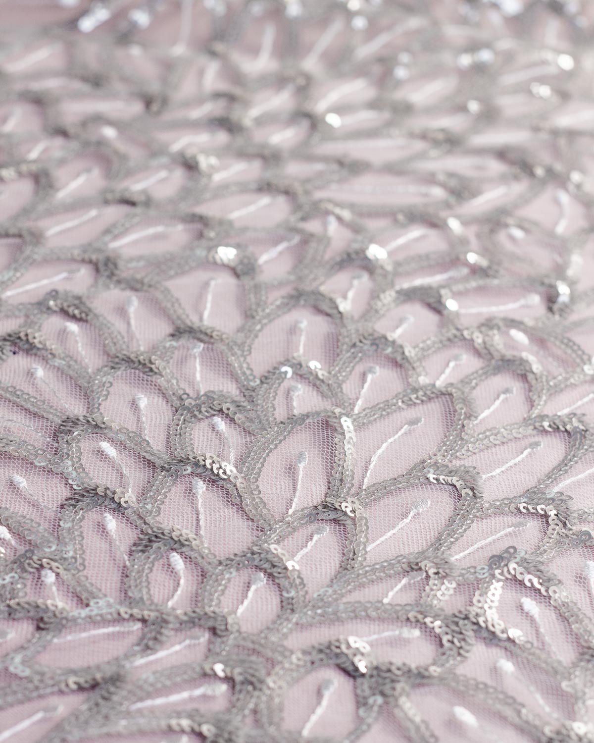A close-up of white tulle mesh fabric decorated with intricate silver sequins and embroidery in a repeating scalloped feather or leaf like pattern.