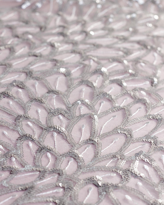 A close-up of white tulle mesh fabric decorated with intricate silver sequins and embroidery in a repeating scalloped feather or leaf like pattern.