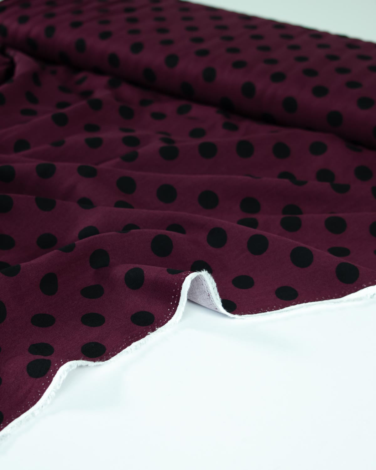 A close-up of a burgundy viscose twill fabric with a black polka dot pattern, draped elegantly on a clean white surface.