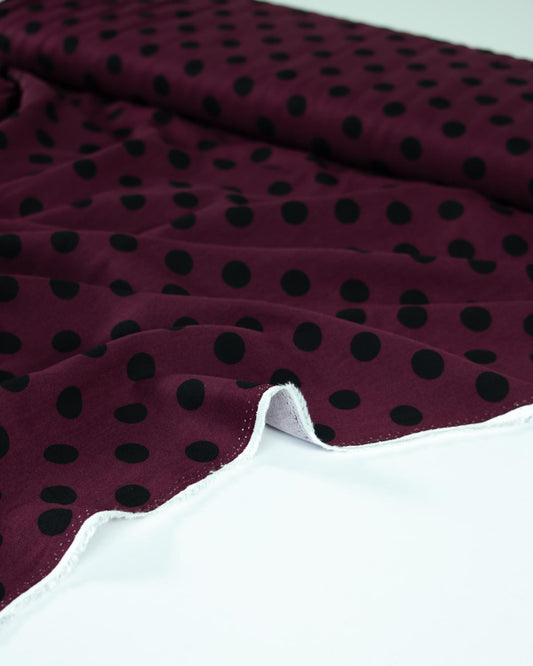 A close-up of a burgundy viscose twill fabric with a black polka dot pattern, draped elegantly on a clean white surface.