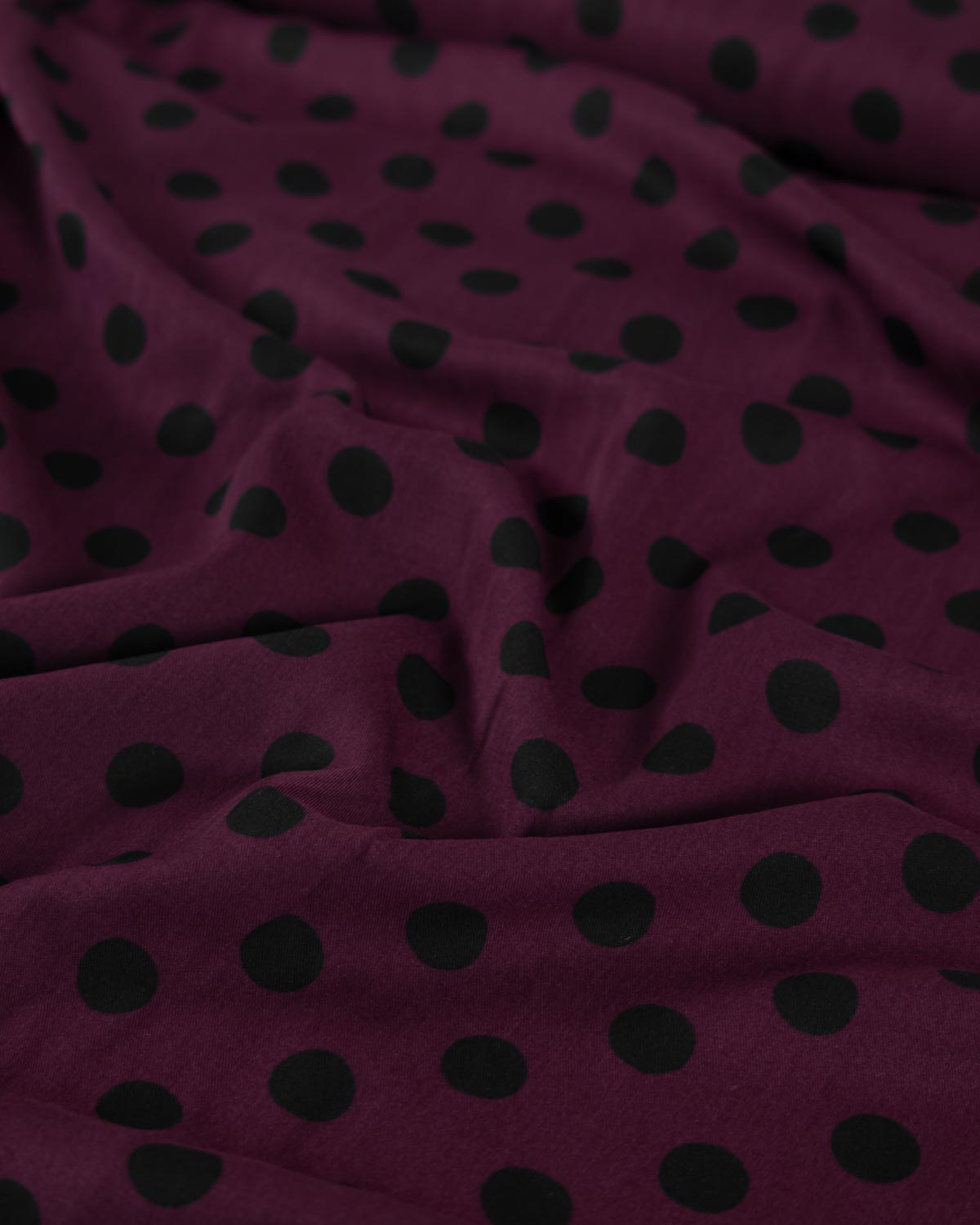 A close-up of draped viscose twill fabric in a deep burgundy or plum purple color with a repeating black polka dot pattern.