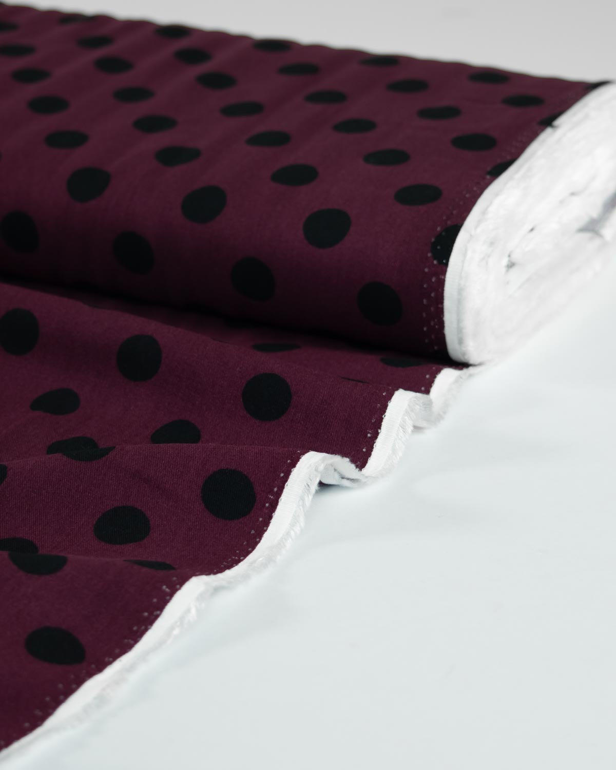 A close-up of a rolled viscose twill fabric with a deep burgundy base and black polka dots, showing a white stitched edge.
