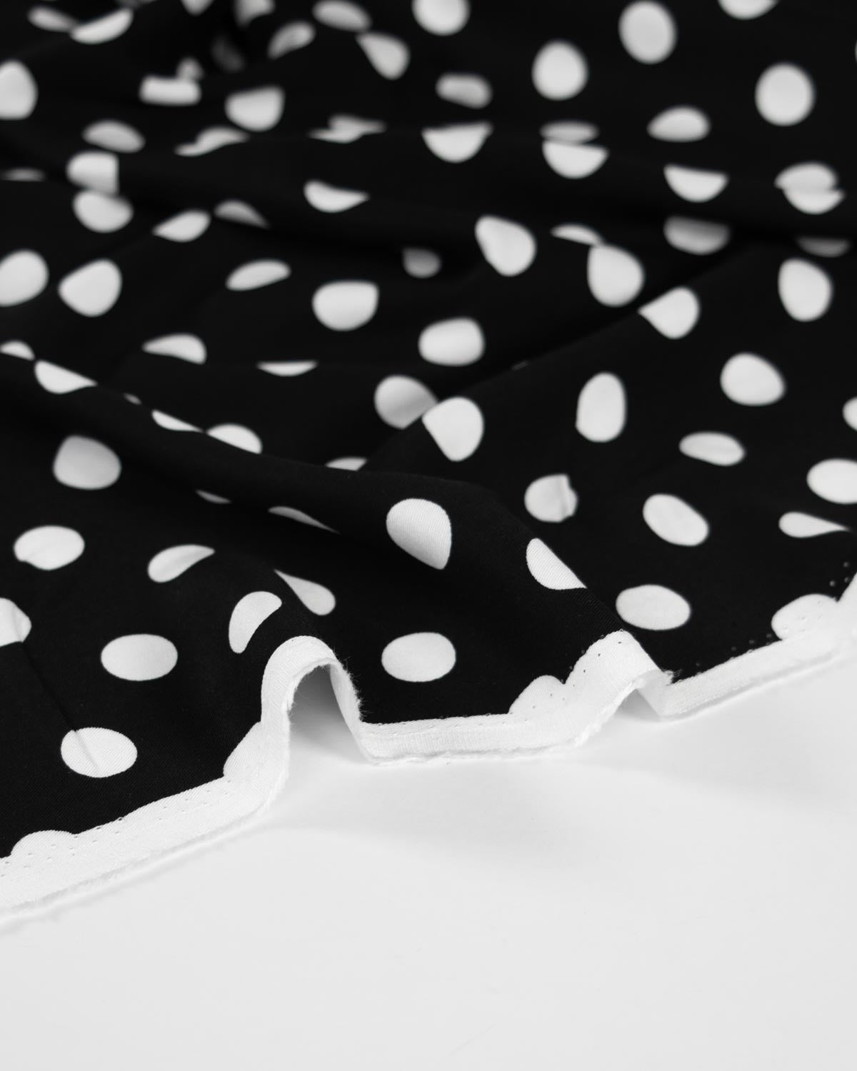 Black viscose twill fabric with a white polka dot pattern and a white scalloped edge, shown in a close-up studio shot.