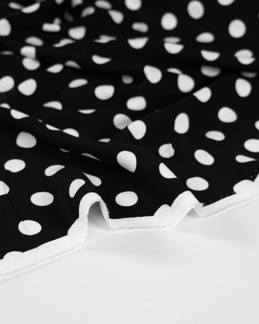 Black viscose twill fabric with a white polka dot pattern and a white scalloped edge, shown in a close-up studio shot.