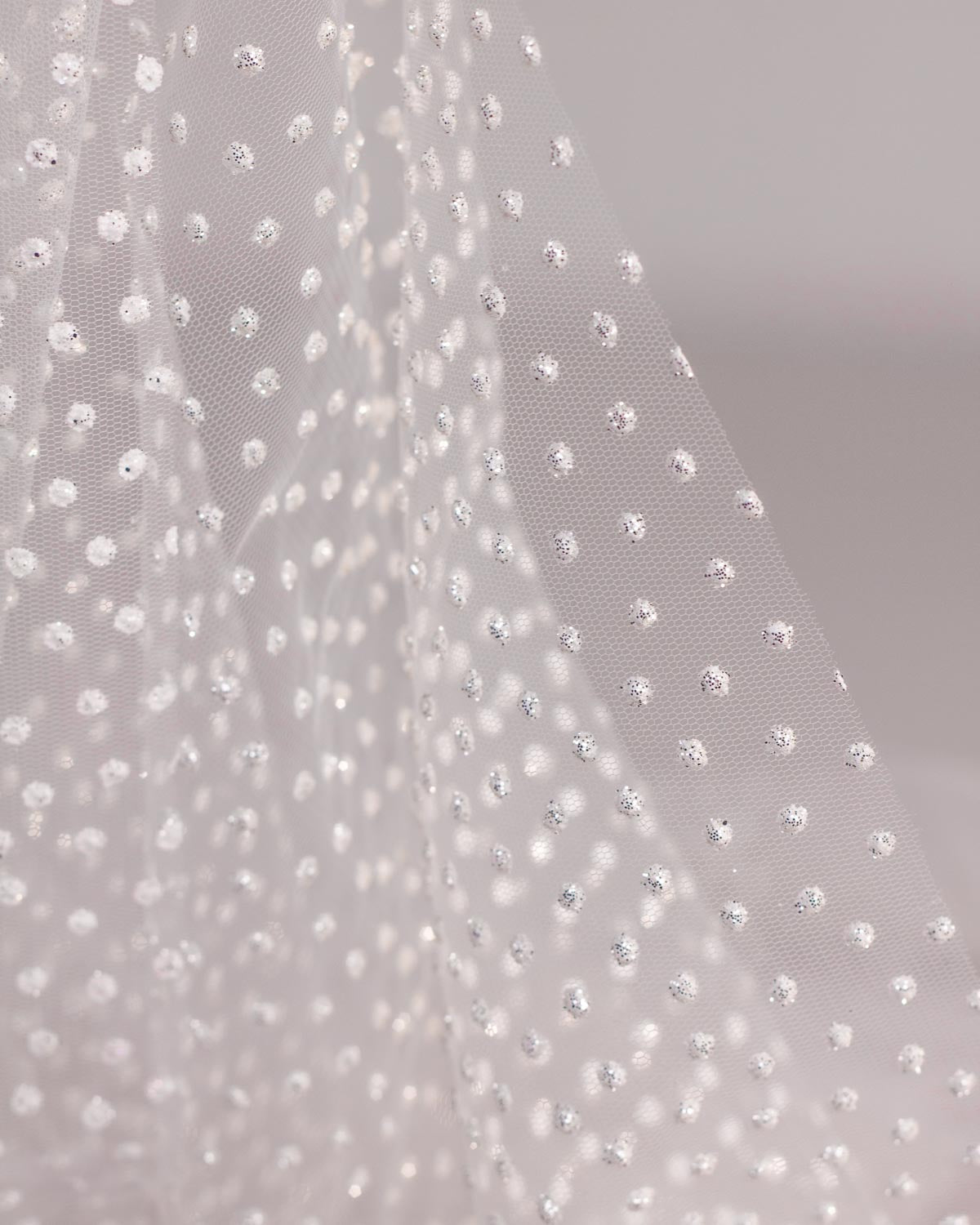 A close-up of draped sheer white polyester tulle fabric decorated with small, evenly spaced white dots with silver glitter. 