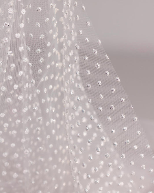 A close-up of draped sheer white polyester tulle fabric decorated with small, evenly spaced white dots with silver glitter. 