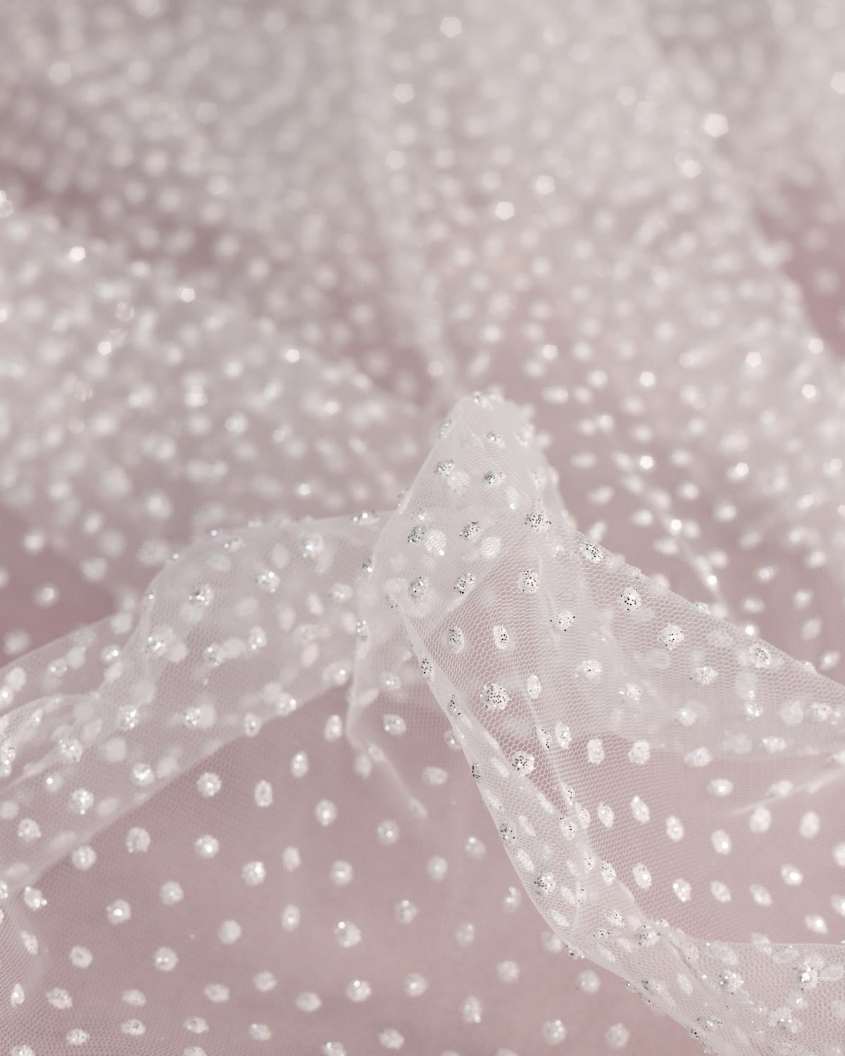 A close-up of a sheer white tulle fabric with a dense pattern of small white dots with silver glitter, draped over a soft pink background.