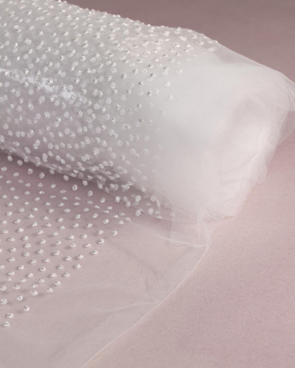 White polyester tulle fabric with dense white dot with silver glitter embellishment, shown rolled out on a soft pink background.